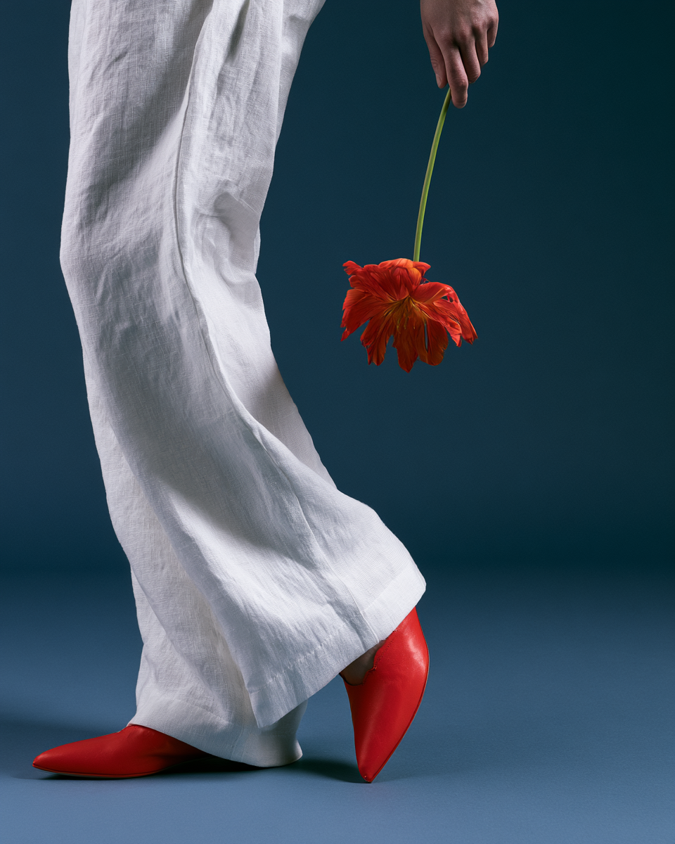 Editorial poster med vita linnebyxor, röda skor och en böjd röd blomma mot djup blå bakgrund.